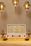 A white pocket temple of Laxmi, Ganesh and Saraswati on a table with decoratives like flowers and diyas with lanterns in the background